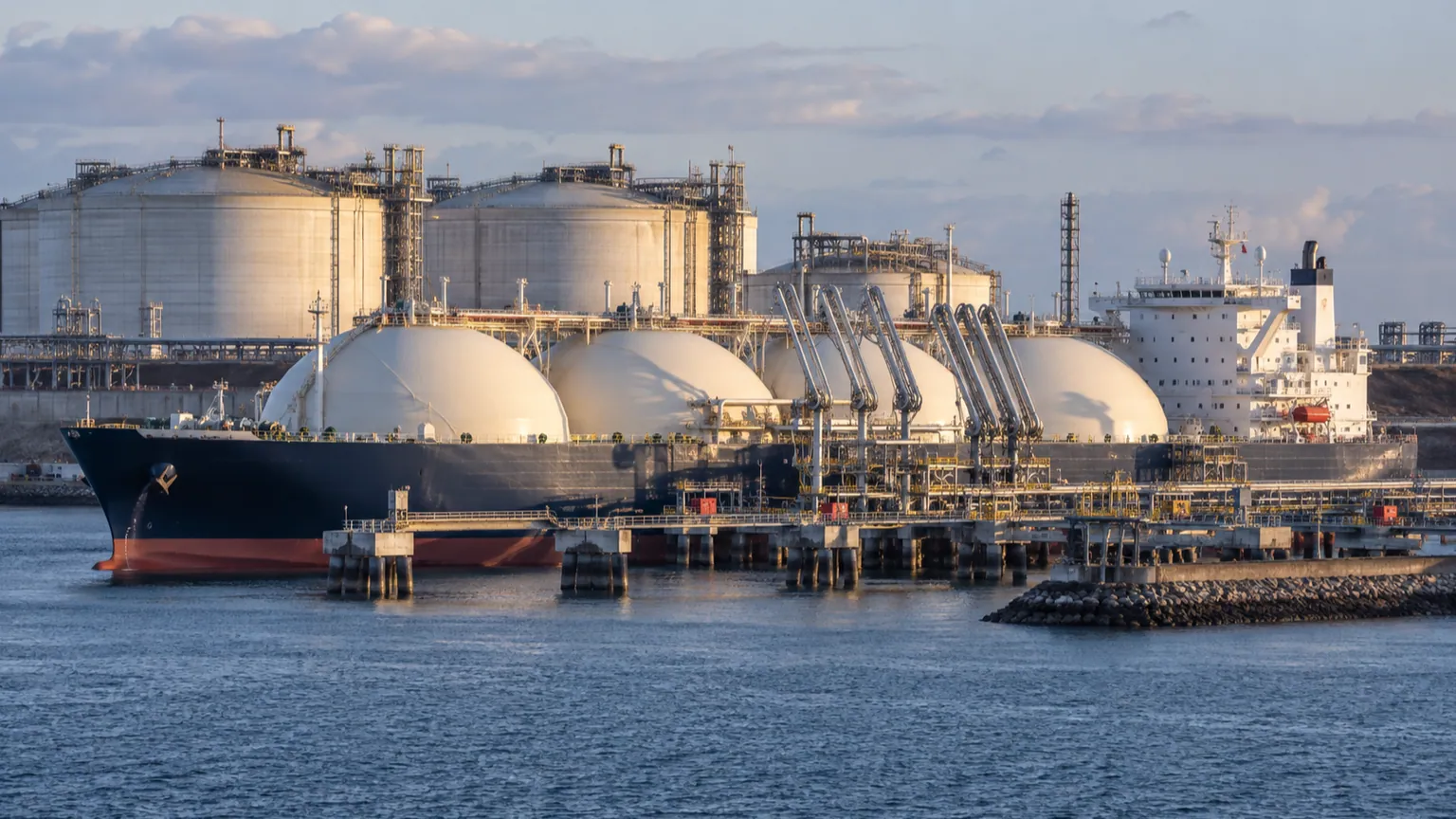 Gaz européen et cargaisons de GNL — méthanier accosté à un terminal, bras de déchargement reliés au navire et réservoirs industriels au bord de la mer.
