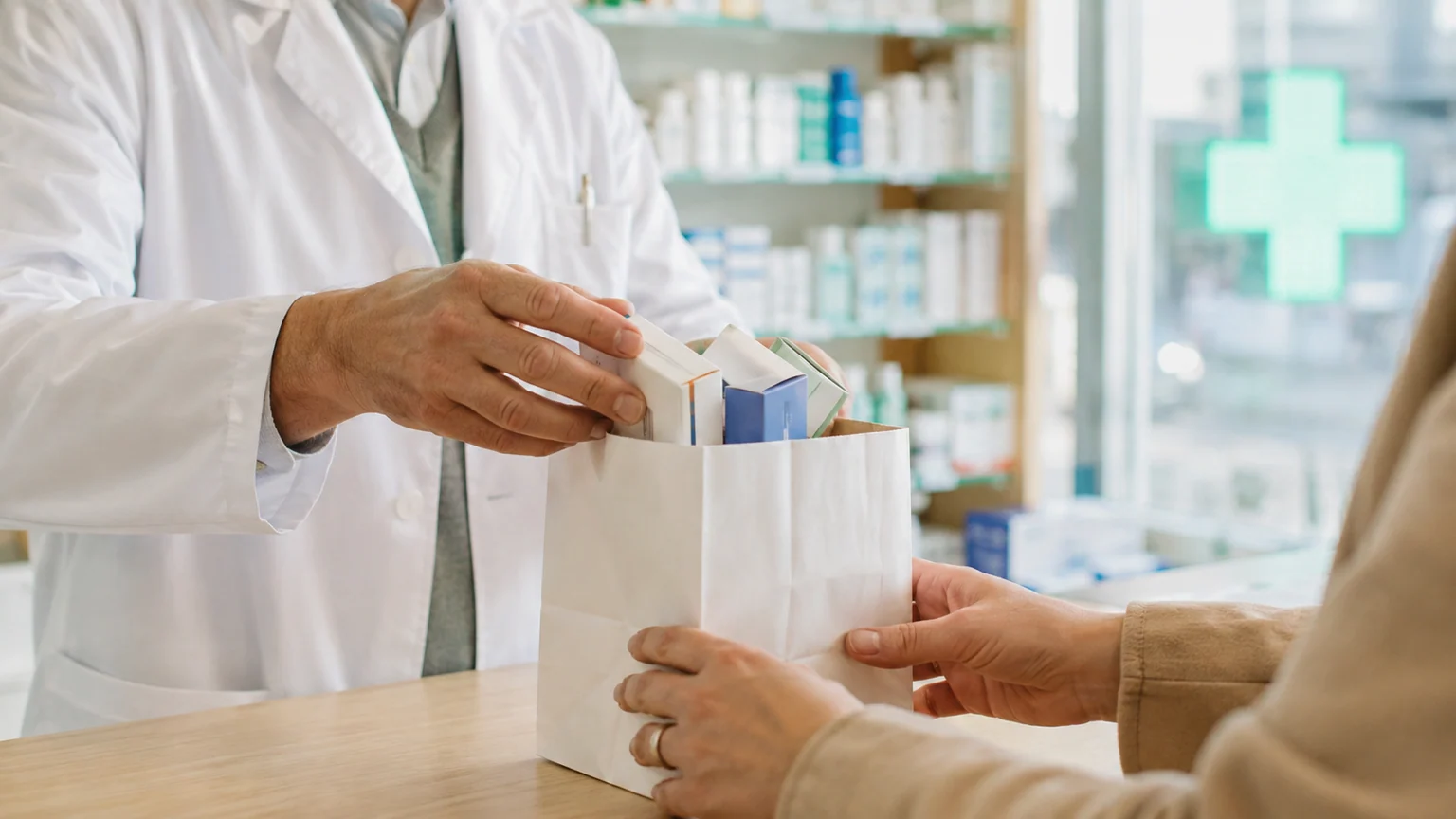 Remboursement des médicaments — mains d’un pharmacien plaçant plusieurs boîtes sans marque dans un sac blanc, devant une croix verte.