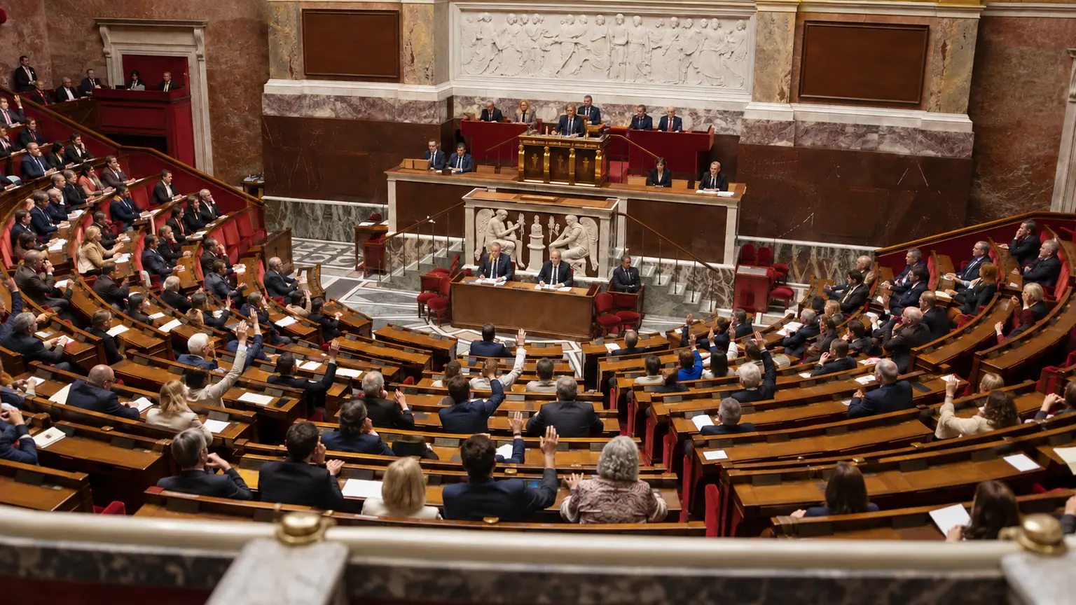 Taxe Zucman rejetée par l’Assemblée — hémicycle français vu depuis les tribunes, pupitres et boîtiers de vote visibles, députés non identifiables levant la main.